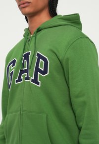 Sweatshirt verde com fecho, feito em tecido macio, destacando um grande logótipo "GAP" em azul-marinho, um bolso frontal e um capuz com cordão.