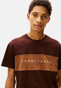 Camiseta de algodón marrón con un panel horizontal beige que muestra el logo "TOMMY JEANS" en puntadas blancas. Cuenta con cuello redondo y mangas cortas.