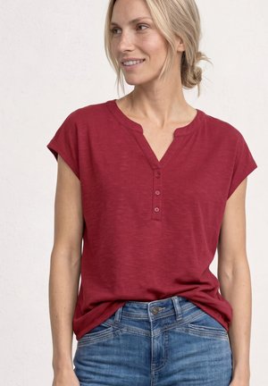 Kvinde iført en rød henley med korte ærmer og knapper, løst stoppet ned i blå denimjeans, som kigger til siden og smiler.