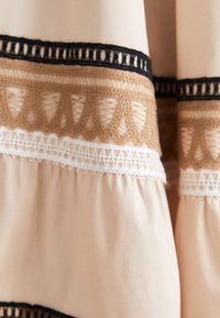 Tissu beige avec des bandes horizontales décoratives de motifs de dentelle complexes en noir, marron et blanc.