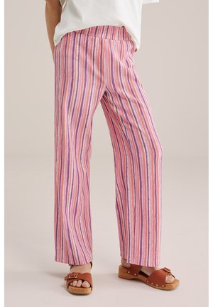 Persona che indossa pantaloni a gamba larga a righe rosa, viola e bianche con sandali in legno marrone e una maglietta bianca, in piedi su uno sfondo chiaro.