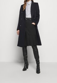 Abrigo de lana azul marino con diseño de doble botonadura, combinado con una camisa de cuadros, una falda negra con pliegues, y botas de cuero negro hasta la rodilla.