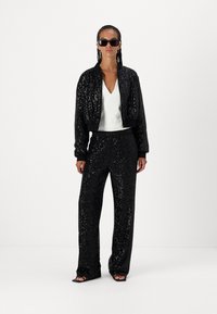 Black sequined bomber jacket with a V-neck white top and matching sequined wide-leg pants; model wears sunglasses and strappy black heels.