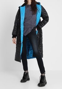 Abrigo largo acolchado en negro y azul con diseño en colcha, llevado sobre una blusa gris con estampado y jeans oscuros, combinado con botas negras de cordones.