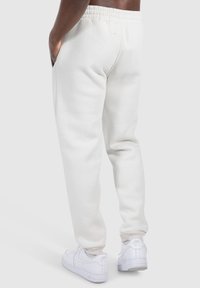 Weiße Baumwoll-Jogginghosen mit elastischem Bund, Seitentaschen und gerippten Bündchen, kombiniert mit weißen Sneakers auf einem einfarbigen Hintergrund.