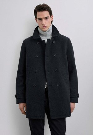 Abrigo de lana gris oscuro de corte cruzado con un alto cuello, bolsillos laterales y mangas abotonadas. Lleva puesto un suéter de cuello alto gris claro.