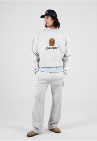 Grijze sweatshirt met een logo en gouden accenten, gecombineerd met bijpassende grijze sweatpants. Gedragen over een lichtblauw, gekraagde shirt. Bruine schoenen.