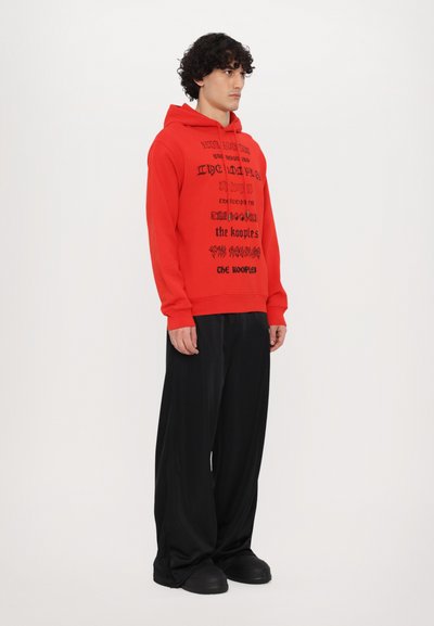 Rode hoodie met zwart grafisch tekstontwerp, gecombineerd met losse zwarte broek en dikke zwarte schoenen. Effen achtergrond, zijprofielweergave.