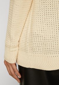 Detailní záběr na ruku vedle krémového pleteného svetru s žebrovanými lemy a spodním okrajem, nošeného přes černé kožené kalhoty.