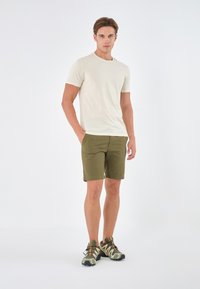 Lys beige t-skjorte og olivengrønne shorts, sammen med sko med camoflasjemønster og gummisåler, stående mot en hvit bakgrunn.
