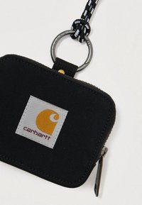 Musta kangaspussukka vetoketjulla, jossa on hopeinen avainrenka ja punottu hihna. Pussukassa on valkoinen pata, jossa on keltainen logo ja punainen teksti.