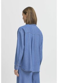 Camicia di jeans azzurro chiaro con maniche lunghe, polsini con bottone e vestibilità morbida. Presenta uno yoke sul retro con dettagli a pieghe e un orlo curvo.