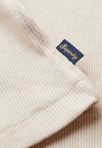 Beige ribbet stof med en blød tekstur; har et blåt mærke mærket "Superdry" i gul kursiv syet ind i hjørnet.