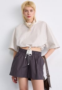 Gestreept, verkort beige top met een koord onderaan, gedragen met donker gestreepte shorts. Gladde textuur, comfortabele pasvorm, aangevuld met een lichte sjaal en tas.