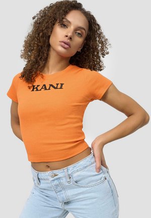 Oransje ribbestrikket crop topp med korte ermer og svart "KANI" tekst, kombinert med lyseblå høytlivsjeans, vist mot en grå bakgrunn.