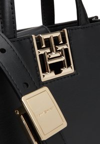 Bolso de mano de cuero negro con un cierre de logotipo geométrico dorado y un detalle de hardware rectangular dorado, con una textura suave y una forma estructurada.