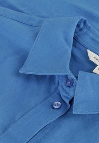 Kortermet blå skjorte laget av lett stoff, med krage som kan kneppes og to blå knapper på lisser.