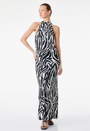 Kobieta stojąca z ręką na biodrze ubrana w bezrękawnikową, czarno-białą sukienkę maxi w zebrę i czarne sandały na paski.