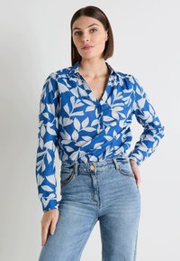 Blusa de botones azul con patrón de hojas blancas; mangas largas, cuello con solapa y tejido ligero. Combinada con jeans de talle alto.