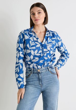 Blusa de botones azul con patrón de hojas blancas; mangas largas, cuello con solapa y tejido ligero. Combinada con jeans de talle alto.