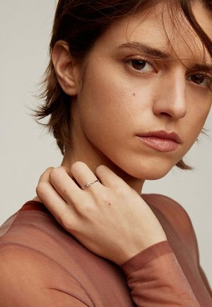 Anillo de plata con una pequeña piedra, llevado en una mano de piel clara. El fondo presenta colores suaves y neutros con una textura discreta.