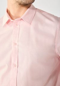 Homme portant une chemise rose clair à boutons avec un col classique, montrant le haut de la poitrine et du cou.