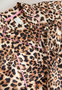 Das Pyjama-Set mit Leopardenmuster besteht aus weichem Stoff, hat braune und schwarze Flecken, pinke Paspeln an Kragen und Tasche sowie einen elastischen Bund.