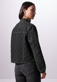 Chaqueta de mezclilla negra acolchada con cuello, puños elásticos y un corte corto. Presenta patrones de diamantes y un acabado limpio en el dobladillo.