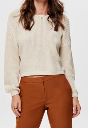 Femme portant un pull en maille côtelée crème à manches longues et un pantalon taille haute de couleur rouille, debout devant un fond uni.