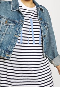 Dżinsowa kurtka w jasnoniebieskim odcieniu, z zapięciem na guziki i kieszeniami na piersi, noszona na paskowanej koszulce w granatowo-białe pasy z niebieskim napisem.