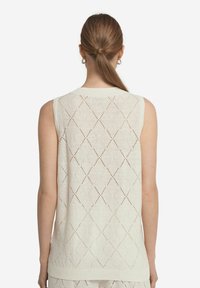 Ärmlös stickad topp i krämfärgad med diamantskärningar, ribbad halsringning och nederkant, mjuk textur som visar en öppen ryggdesign.