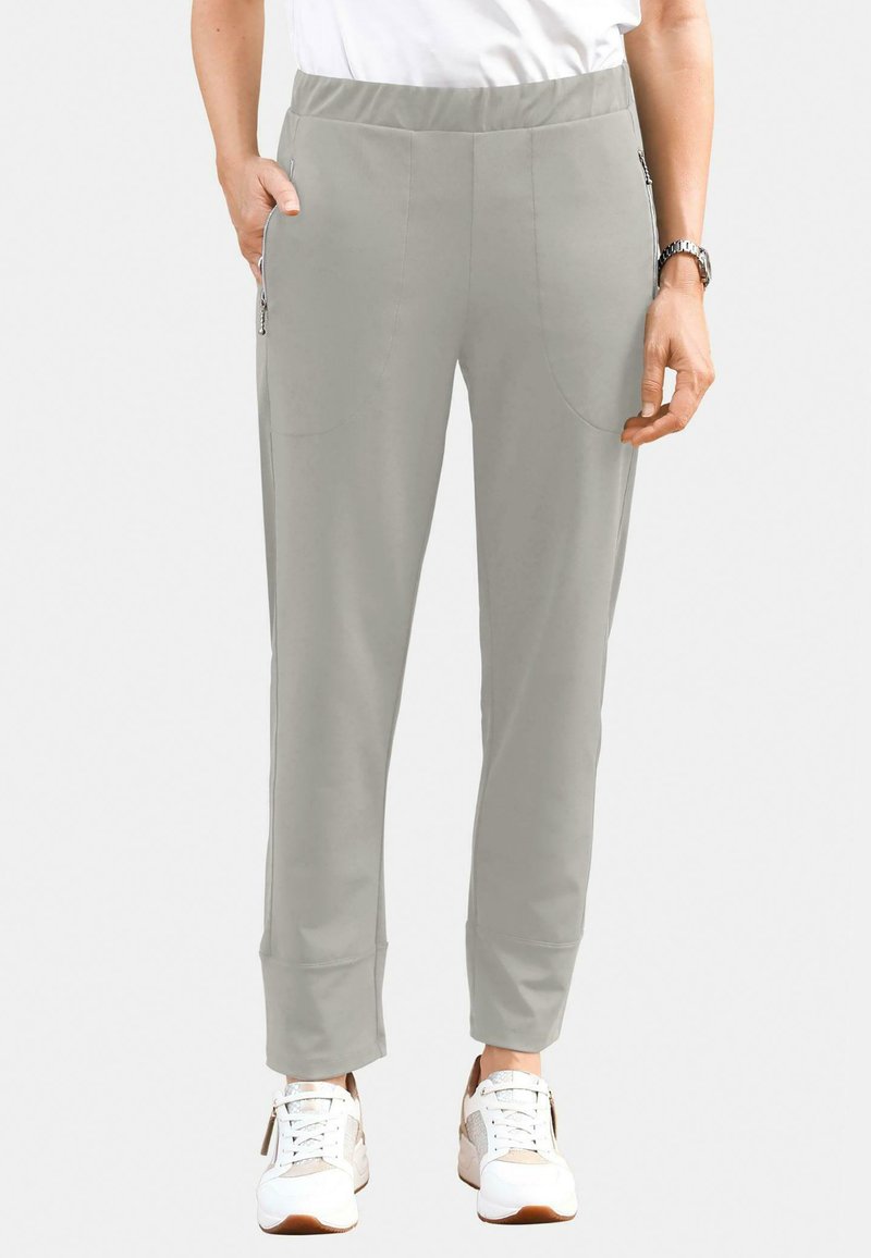 Helle graue Hose mit elastischem Bund, seitlichen Reißverschlusstaschen, schlanker Passform und schmal zulaufenden Knöcheln, kombiniert mit weißen Sneakers.