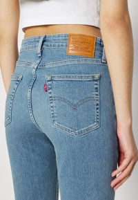 Jeans de denim azul claro con un corte de cintura alta, que presentan un parche de cuero en la parte posterior, dos bolsillos y detalles de costura sutiles.