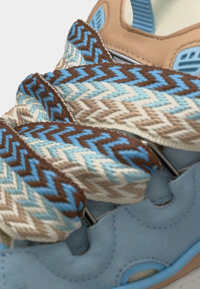 Gros plan sur une basket bleu clair avec des lacets en tissu épais aux motifs en zigzag marron, beige et bleu, noués en haut.