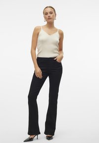 Zwarte flared jeans gecombineerd met een crèmewitte geribbelde tanktop. De top heeft dunne bandjes en een V-halsontwerp. Het model staat met de handen op de heupen.