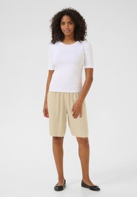 Haut blanc à manches courtes en côte, shorts beige jusqu'aux genoux et ballerines noires. La tenue présente une coupe décontractée et confortable avec une texture lisse.