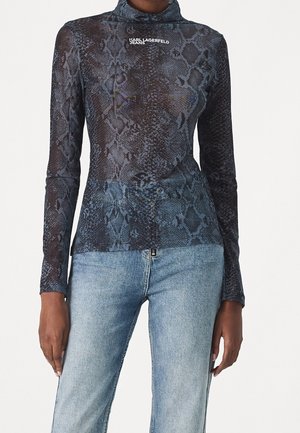 Femme portant un haut transparent à manches longues avec un imprimé serpent bleu-noir et un jean taille haute bleu clair, debout les bras détendus.