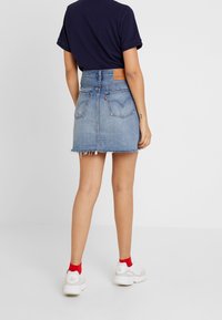 Personne portant une chemise bleue marine, une jupe en jean à ourlet effiloché avec des poches arrière, des chaussettes rouges aux chevilles et des baskets blanches, debout sur un fond uni.