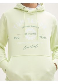 Sudadera verde clara con bolsillo frontal, cordón y texto impreso en blanco que incluye detalles de la marca y calidad. Tela suave y texturizada.