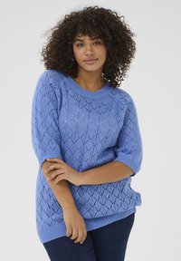 Un pull en tricot bleu clair présentant un motif en losange, des manches courtes et des bords côtelés, assorti à un jean foncé.