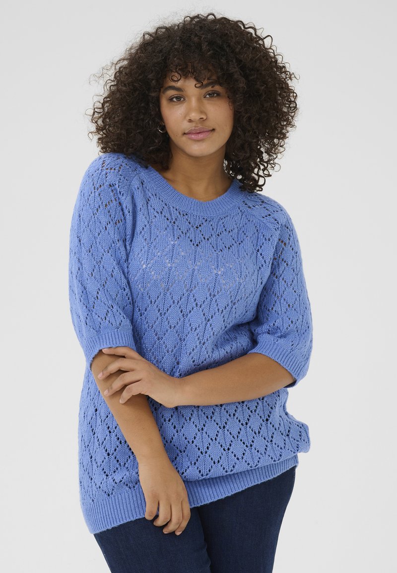 Un pull en tricot bleu clair présentant un motif en losange, des manches courtes et des bords côtelés, assorti à un jean foncé.