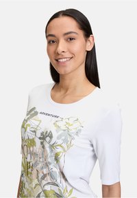 Hvid T-shirt med korte ærmer, prydet med et blomstermotiv i grønne og gule nuancer, og ordet "ADVENTURE" ovenfor grafikdesignet.