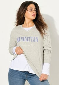 Maglietta a maniche lunghe grigio chiaro con scollatura a V, con la scritta "MANHATTAN" stampata in blu e una sotto-maglia bianca che spunta dall'orlo. Tessuto morbido, vestibilità casual.