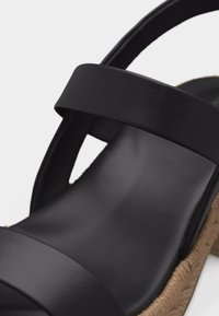 Gros plan d'une sandale noire avec deux larges brides et une semelle beige texturée, montrant l'avant et la zone de la cheville.