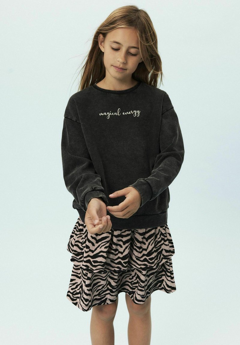 Sweatshirt gris foncé avec le texte "énergie magique", associé à une jupe en couches à rayures zébrées roses, mettant en avant une coupe décontractée et une texture douce.
