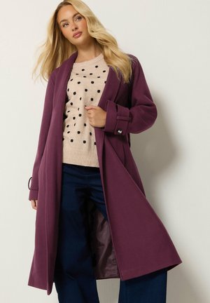 Femme portant un long manteau violet foncé sur un pull beige à pois noirs et un pantalon bleu foncé, debout devant un fond clair uni.