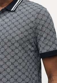 Κοντινό πλάνο ενός γκρι πόλο πουκάμισου με σκούρο κυκλικό μοτίβο, σκούρο μπλε γιακά με λευκές ρίγες και σκούρη μπλε επένδυση στα μανίκια στο χέρι ενός ατόμου.