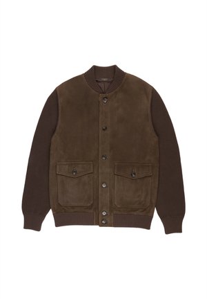 Man draagt een donkerbruine jas over een bruin overhemd, beige broek met een geweven riem en crèmekleurige sneakers, staand tegen een effen achtergrond.