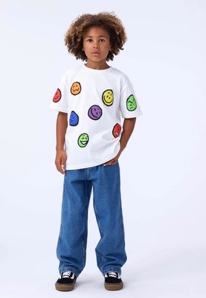 Weißes T-Shirt mit kurzen Ärmeln und bunten Smiley-Gesicht-Grafiken in Rot, Grün, Gelb, Blau und Lila. Kombiniert mit lockeren blauen Jeans und schwarzen Schuhen.