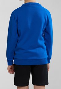 Felpa blu con colletto e polsini a costine, dal taglio rilassato e tessuto testurizzato. Indossata con pantaloni corti neri, con dettagli di cucitura visibili.